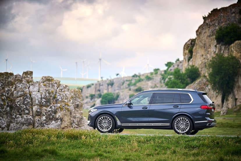 Dark blue BMW X7 SUV parked on a roadside with rocky cliffs and wind turbines under a cloudy sky.