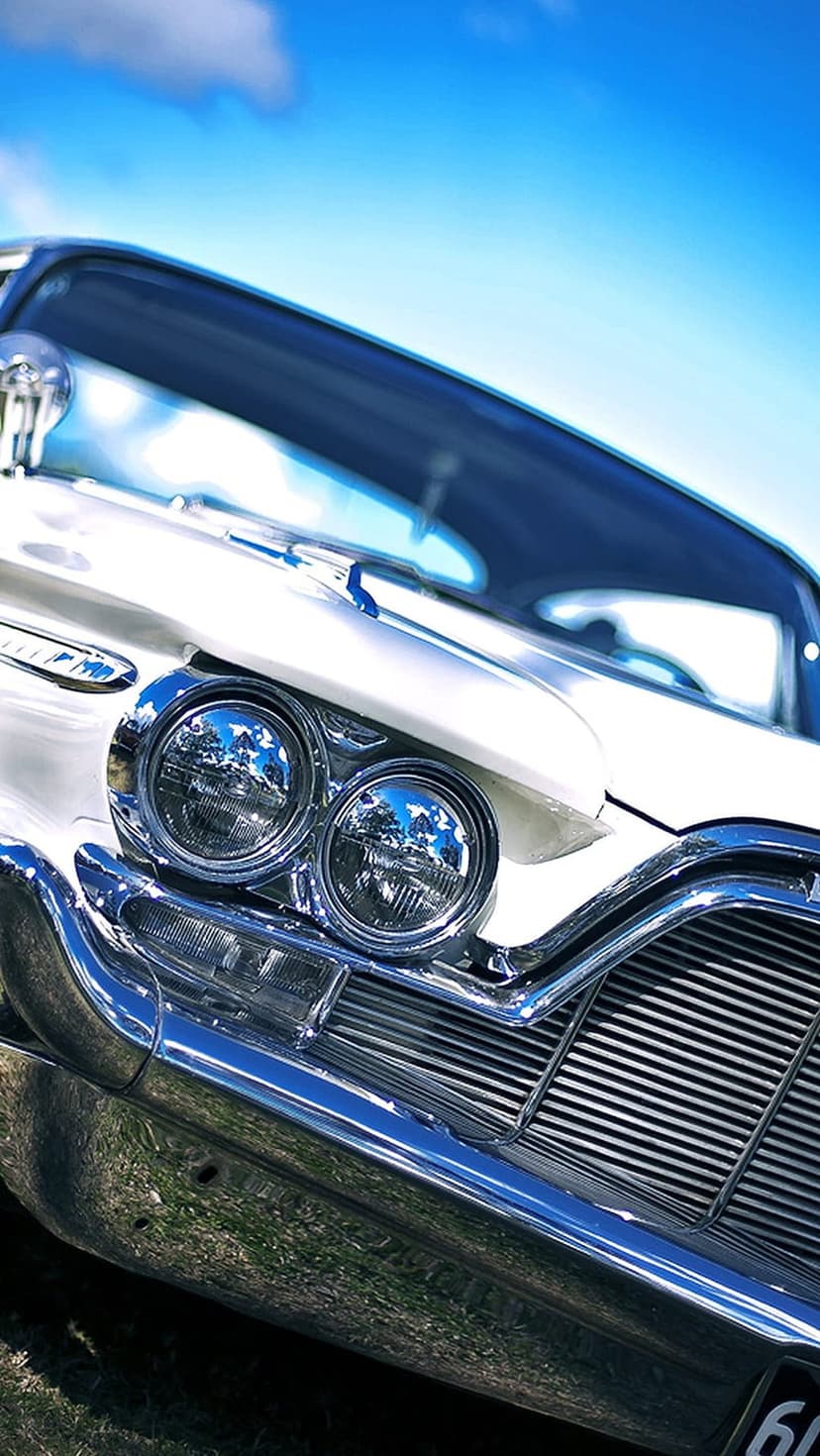 White vintage car front with dual round headlights and a chrome grille reflecting a blue sky and clouds.