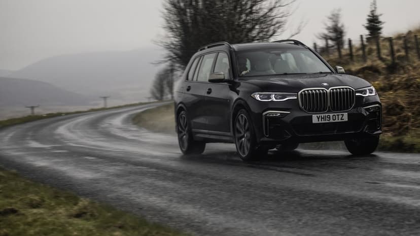 Black BMW X7 SUV driving on a winding wet road through misty hills under a grey overcast sky.