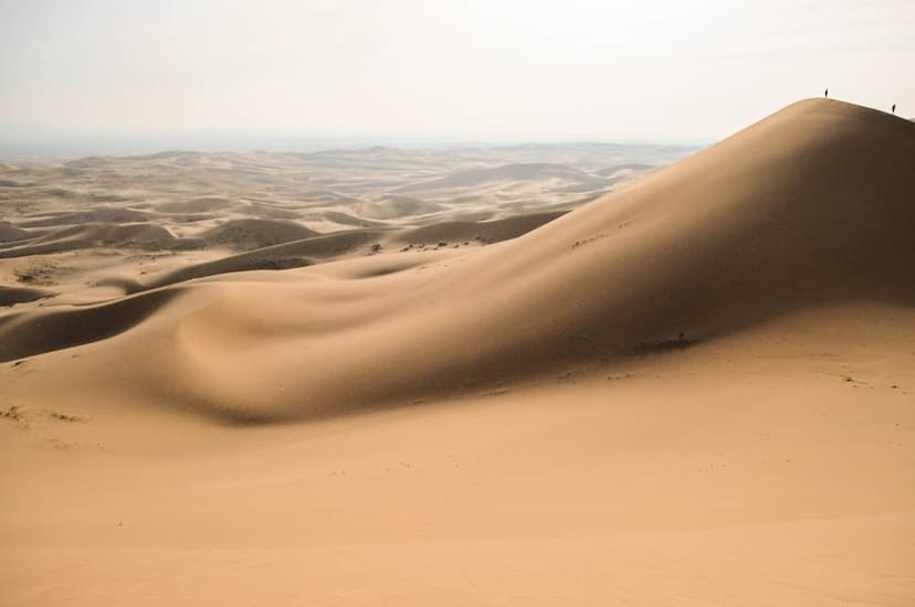 Undulating golden sand dunes lead to two tiny human figures standing on a distant ridge under a soft hazy sky.