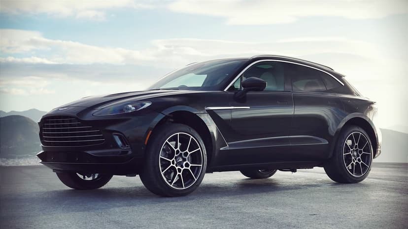 Black Aston Martin DBX SUV parked on a mountain overlook under a cloudy sky with reflective bodywork.