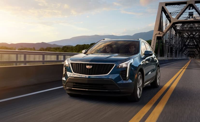 Blue Cadillac SUV driving on a steel truss bridge during a golden sunset with mountains in the background.