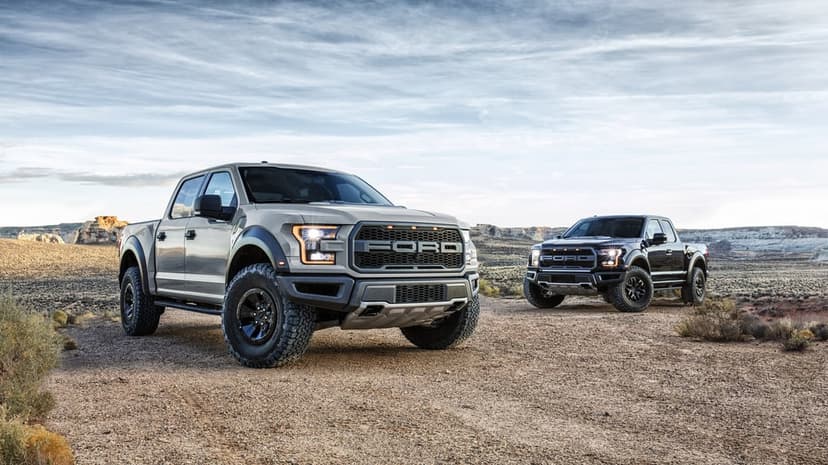 Gray and black Ford F-150 Raptor trucks parked on a dirt trail in a vast desert landscape under a cloudy sky.