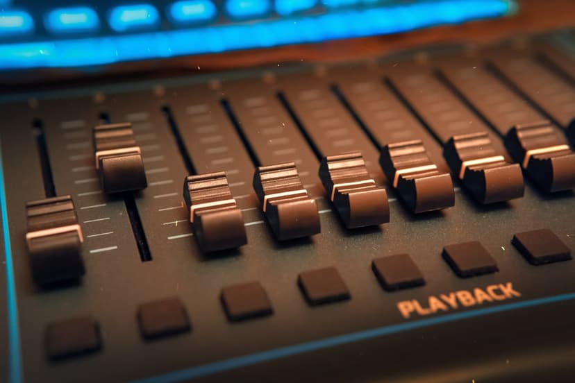 Audio mixing faders in varied positions on a dark slate console with illuminated playback buttons and blue trim.