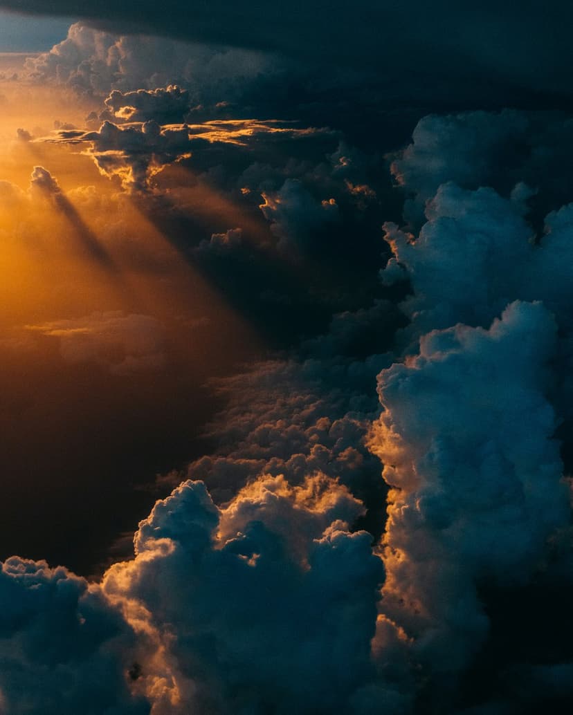 Golden sun rays breaking through dark storm clouds during a high-altitude aerial sunset over a vaporous sky.