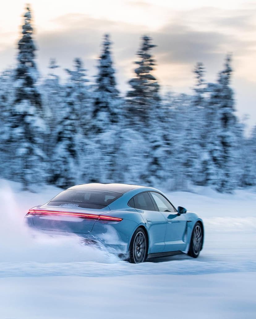 Blue Porsche Taycan drifting on a snowy road through a pine forest with illuminated taillights and snow spray.
