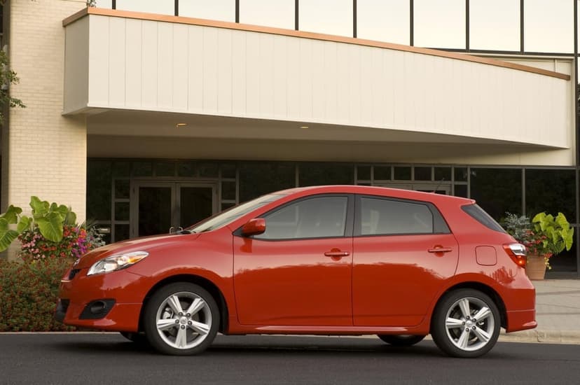 Side profile of a red Toyota Matrix hatchback parked on asphalt in front of a modern white building exterior.