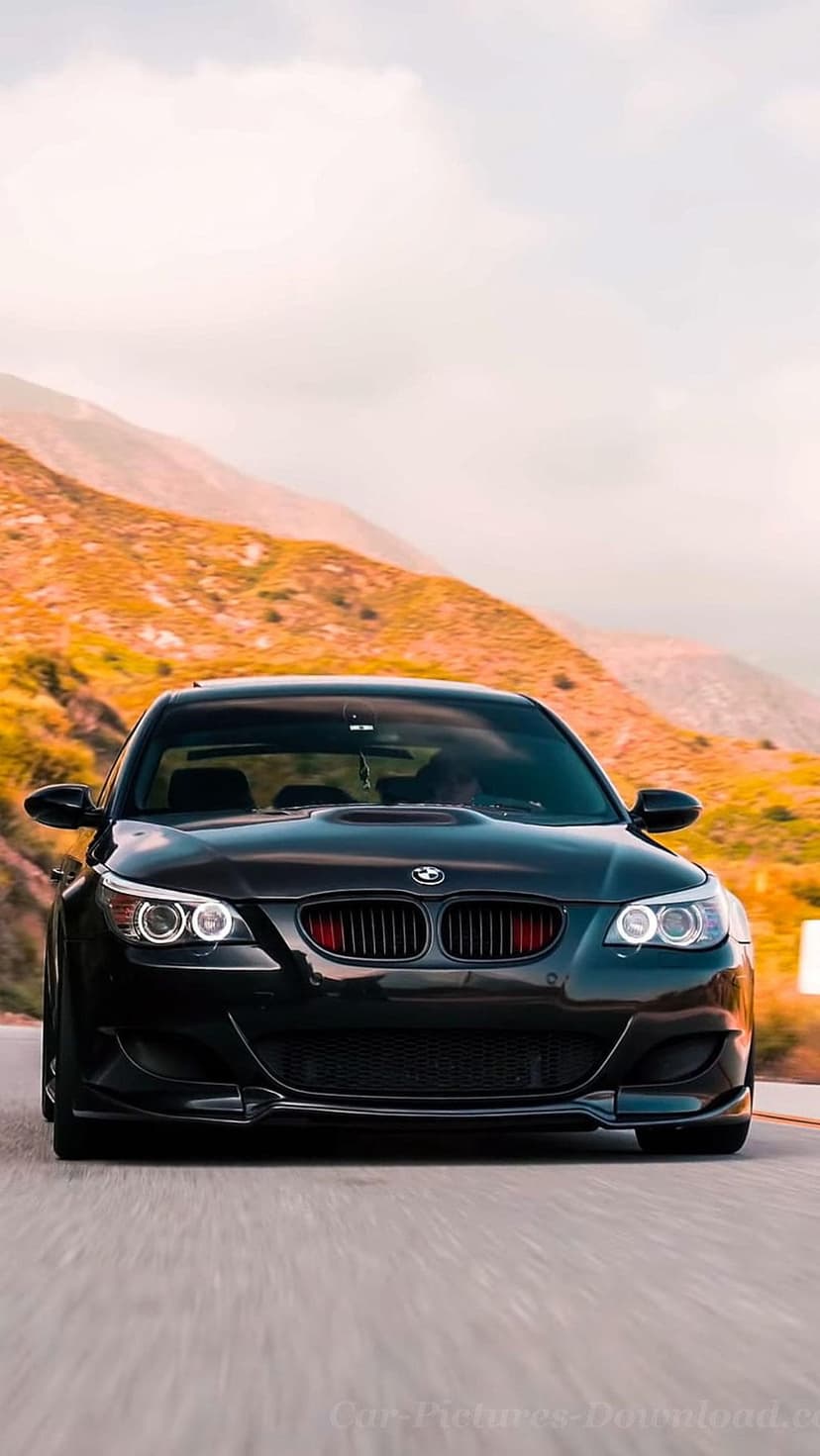 Black BMW M5 sedan with red grille accents driving on a winding road toward golden sunlit mountains.