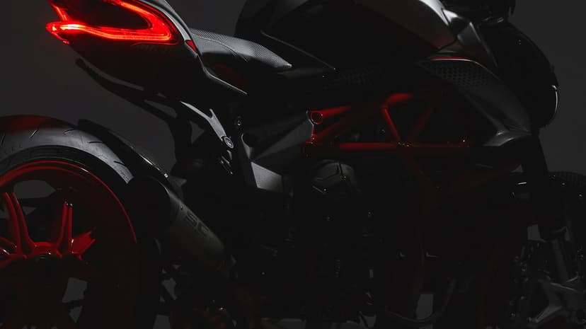 Rear view of a matte black motorcycle with a glowing red taillight and red trellis frame in low light.