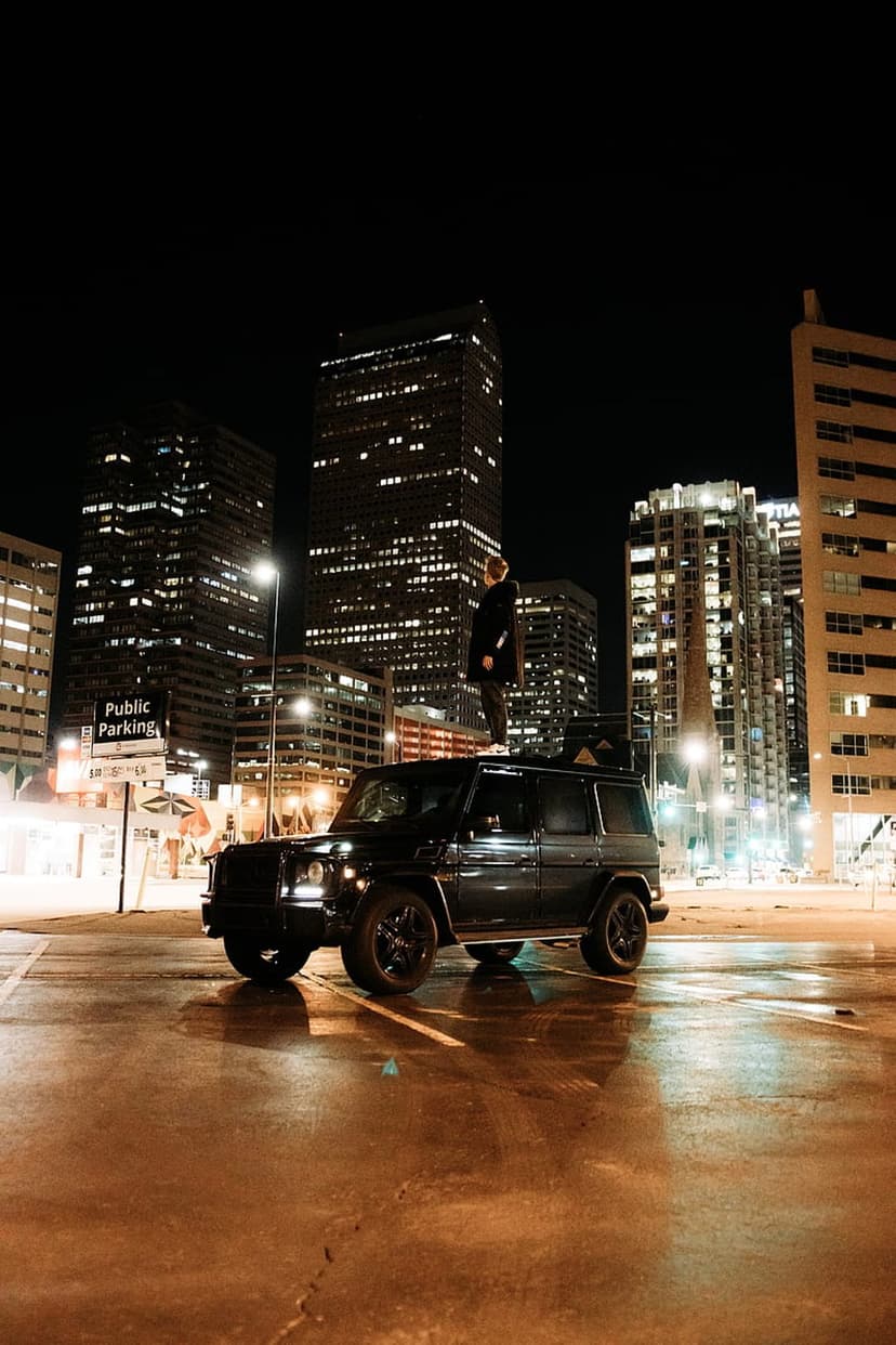 Man standing on a black SUV roof on a wet city street at night with glowing skyscrapers in the background.