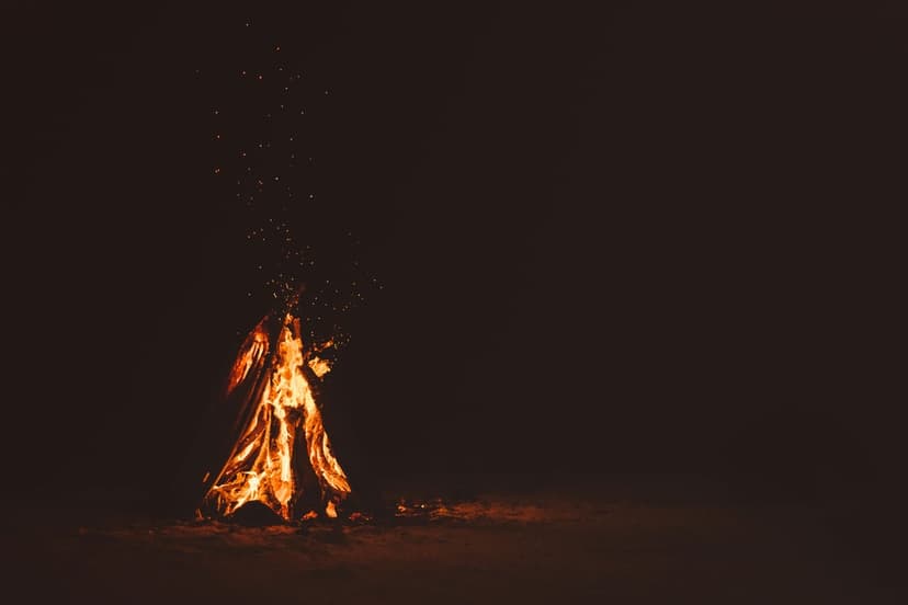 Vivid orange campfire flames and swirling sparks rise from glowing embers against a dark night sky background.