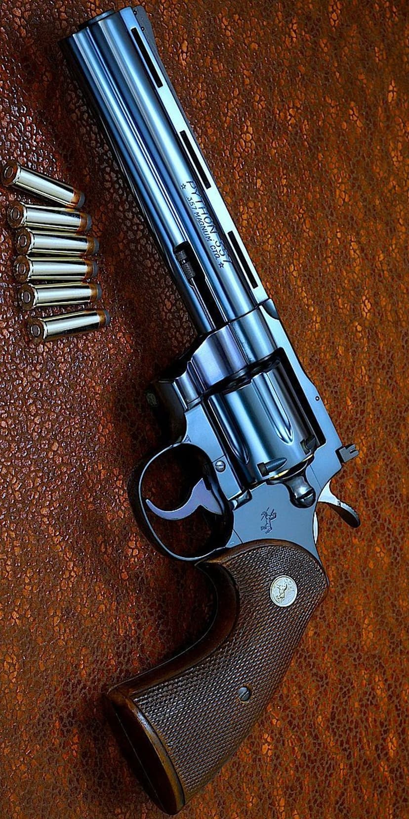 Colt Python revolver and brass rounds sit on a dark brown textured surface in a close-up vertical shot.