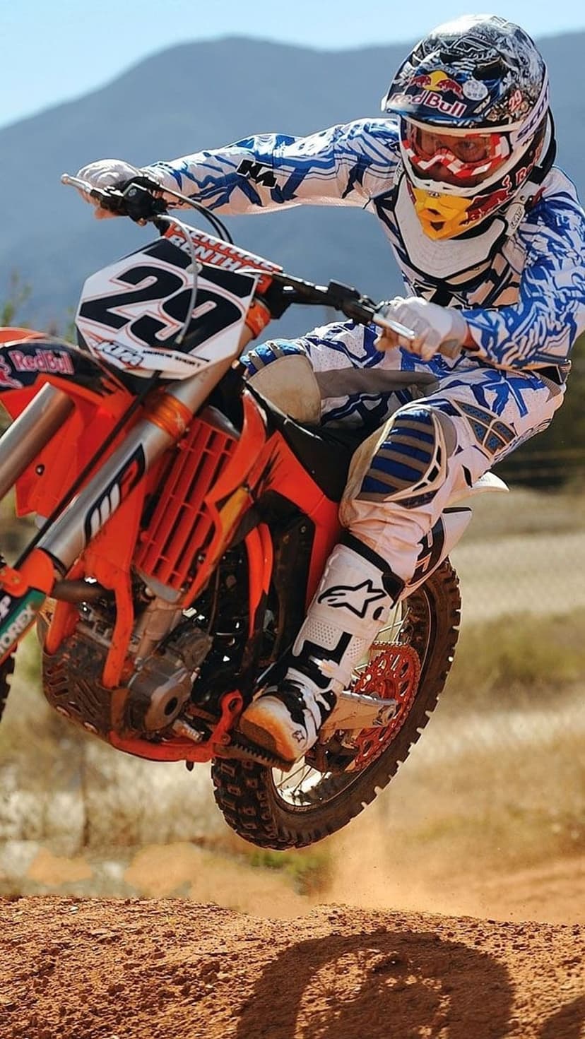 Motocross rider in a blue suit jumps an orange dirt bike mid-air against a blurred mountain landscape.