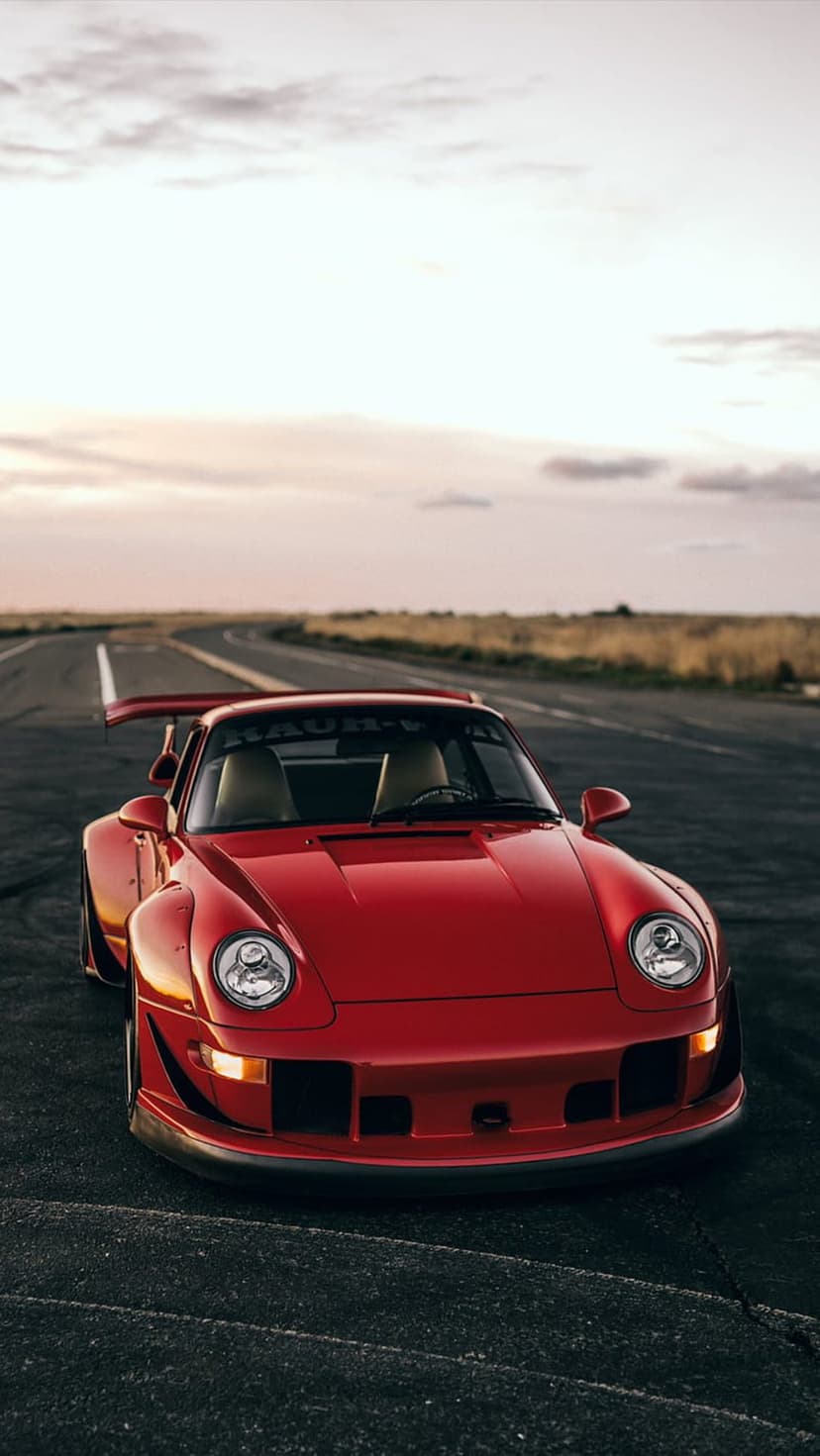 Red Porsche 911 Turbo sports car on a desert road under a warm sunset sky in a low-angle vertical photograph.