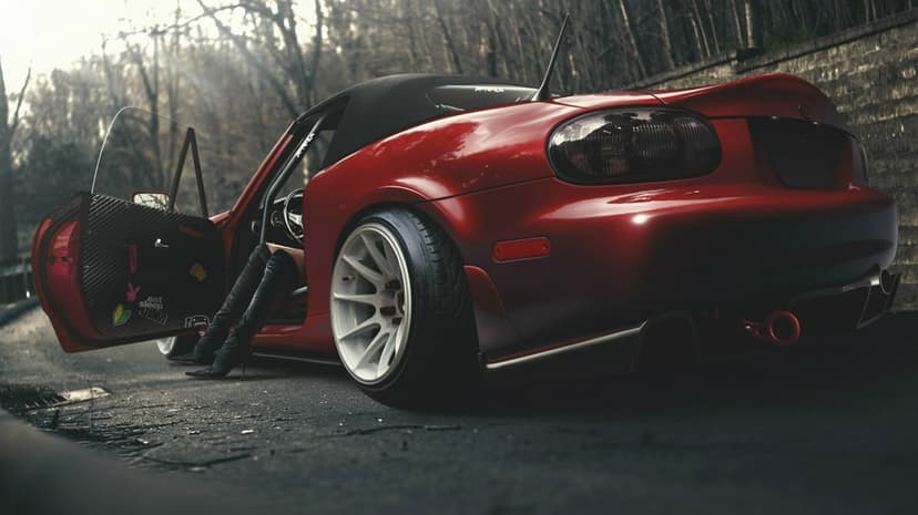 Red Mazda MX-5 Miata with white wheels and open door parked on a misty road near forest trees.