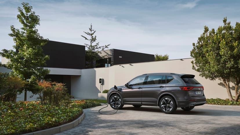 Grey electric SUV charging at a modern house driveway with green trees under a clear blue sky in bright daylight.