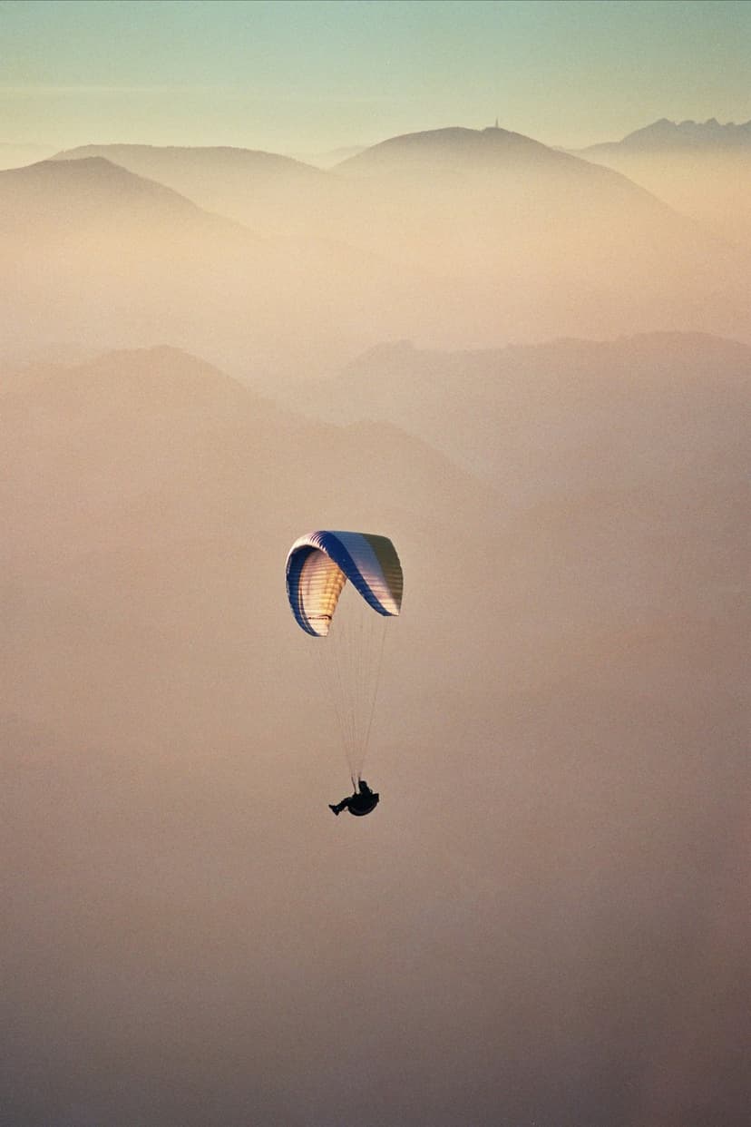 Paraglider with an illuminated canopy soaring above misty mountain peaks during a vibrant golden hour sunset.