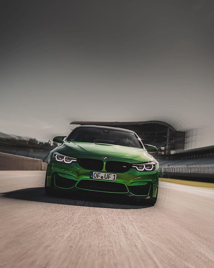 Green BMW M4 F82 coupe speeds on a racetrack with motion blur against a cloudy sky and blurred grandstands.