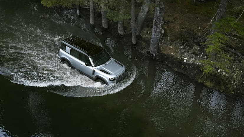 Silver Land Rover Defender wading through a dark forest river, creating white water splashes and ripples.