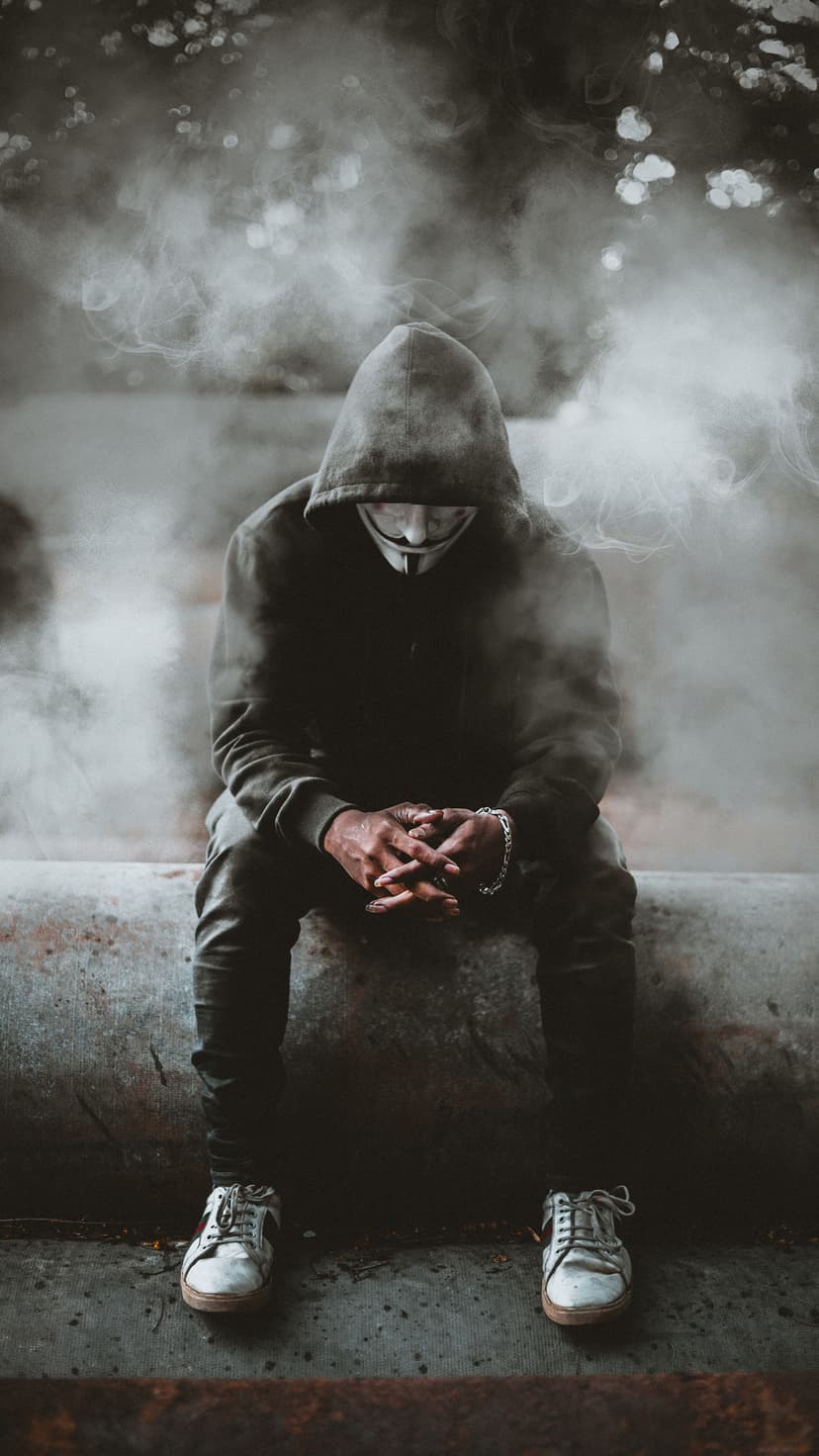 Masked figure in a black hoodie sits on the floor amidst swirling white smoke in a dark, gritty urban setting.