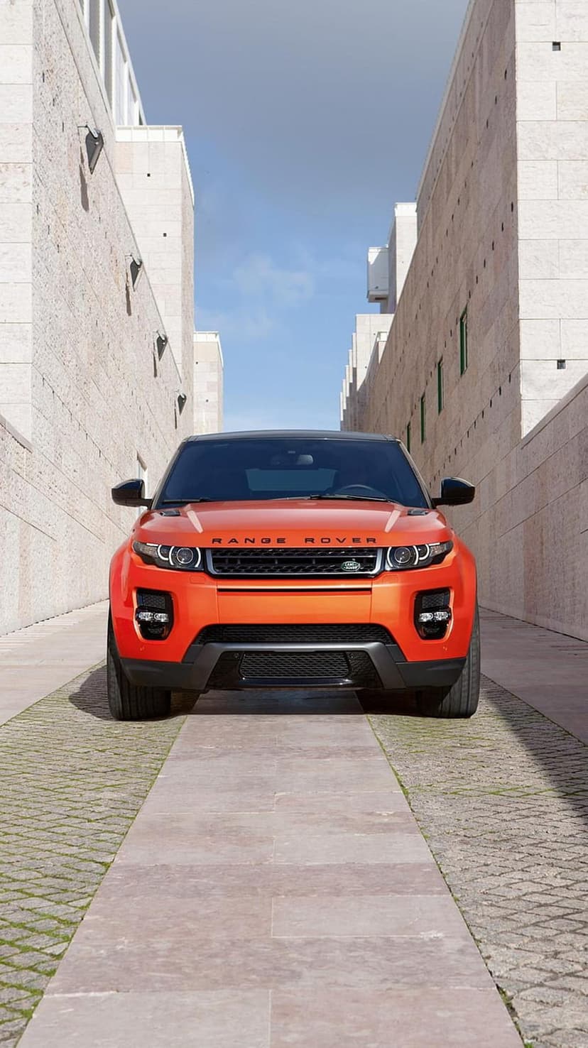 Orange Range Rover Evoque parked on a stone walkway between modern minimalist buildings under a clear blue sky.