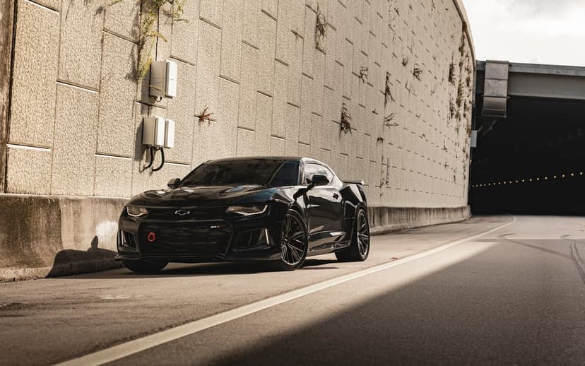 Black Chevrolet Camaro ZL1 parked on a road next to a textured concrete wall under bright golden sunlight.