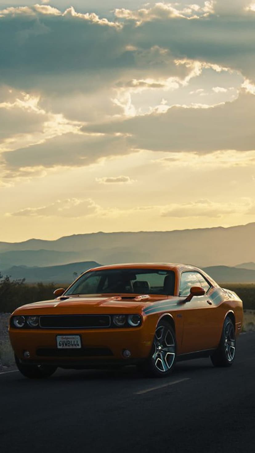 Orange Dodge Challenger with black stripes driving on a desert road under a colorful sunset sky with mountains.