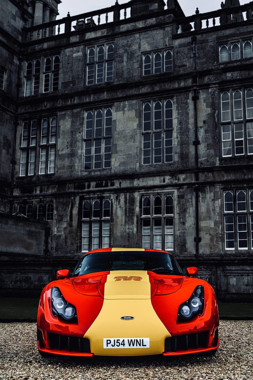 Red TVR Sagaris sports car parked on gravel in front of a massive weathered stone castle with gothic windows.