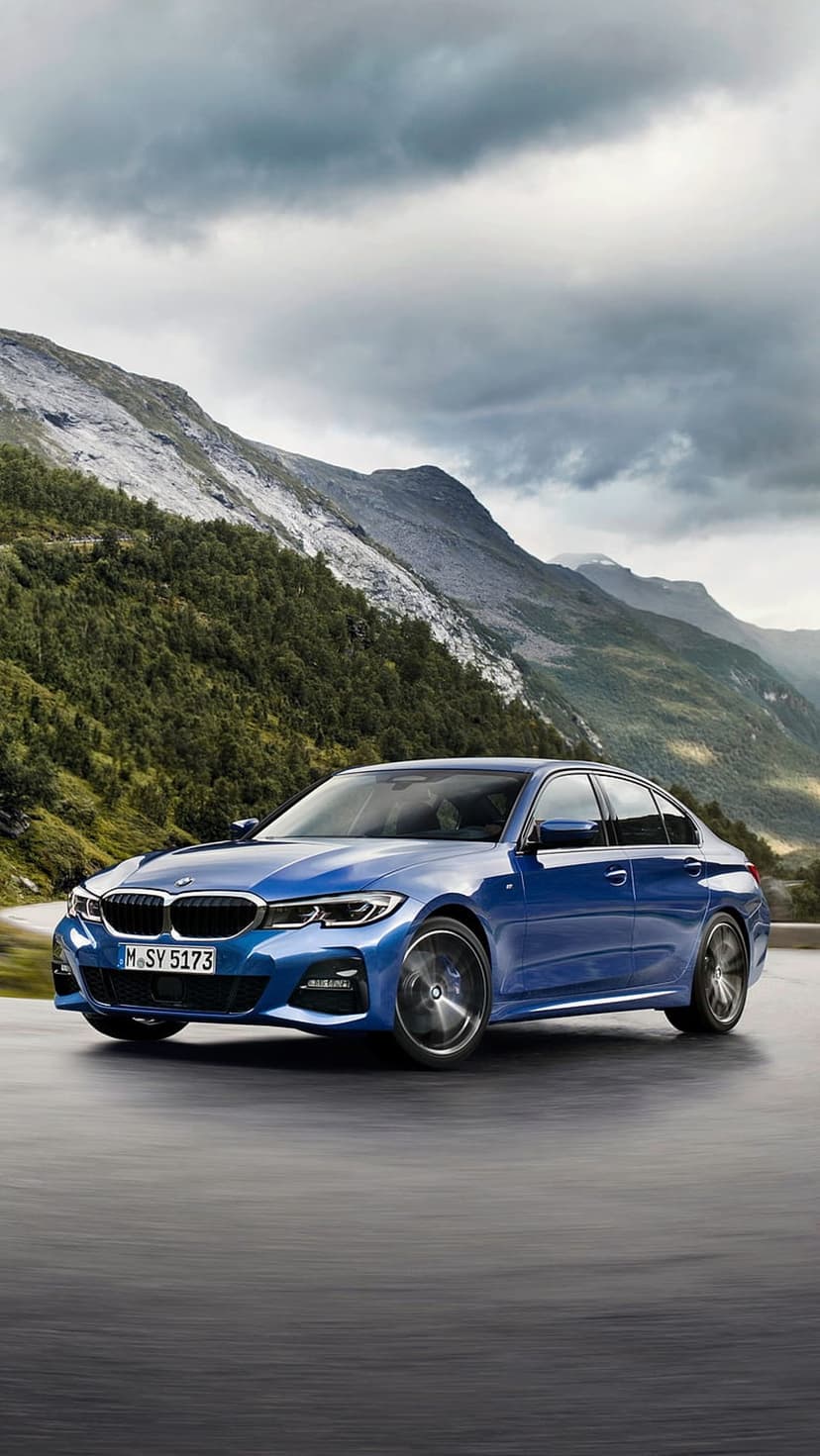 Blue BMW 3 Series sedan driving on a curved mountain road with evergreen trees and rocky peaks in the background.