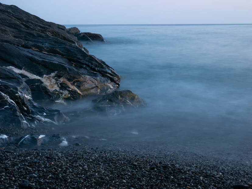 Jagged dark rocks and smooth pebbles meet a misty, long-exposure ocean shore under a soft twilight haze.