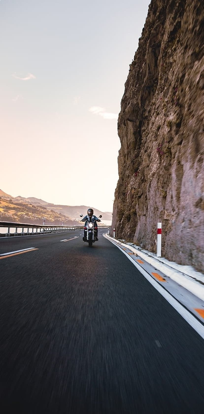 Motorcyclist riding along a curving mountain road overlooking a lake during a golden sunset with rocky cliffs.