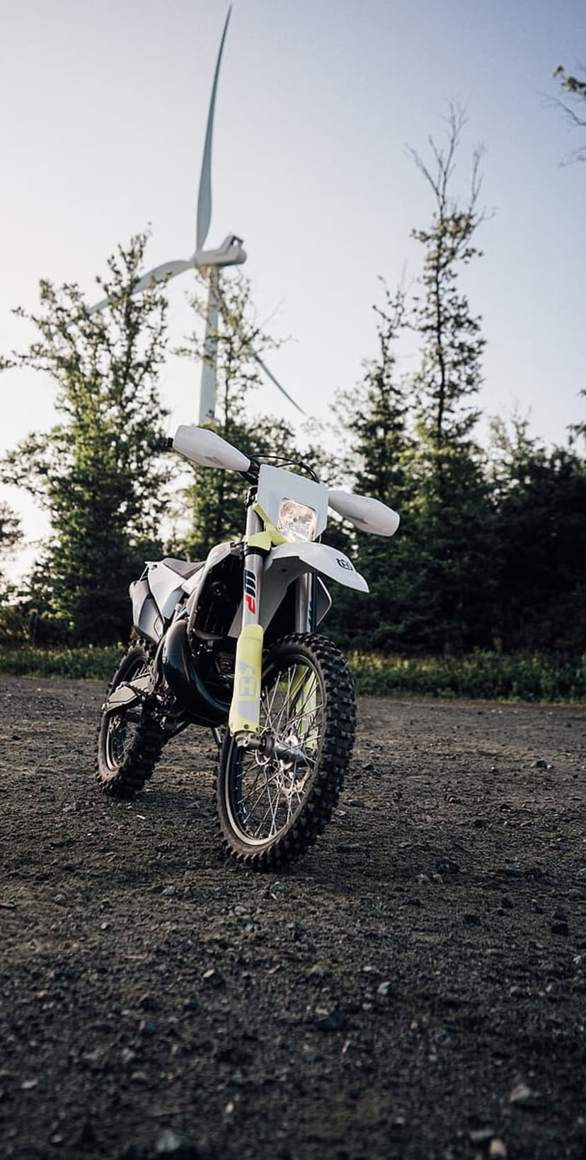 Dirt bike with yellow suspension parked on gravel before a tall wind turbine under a clear bright sky.