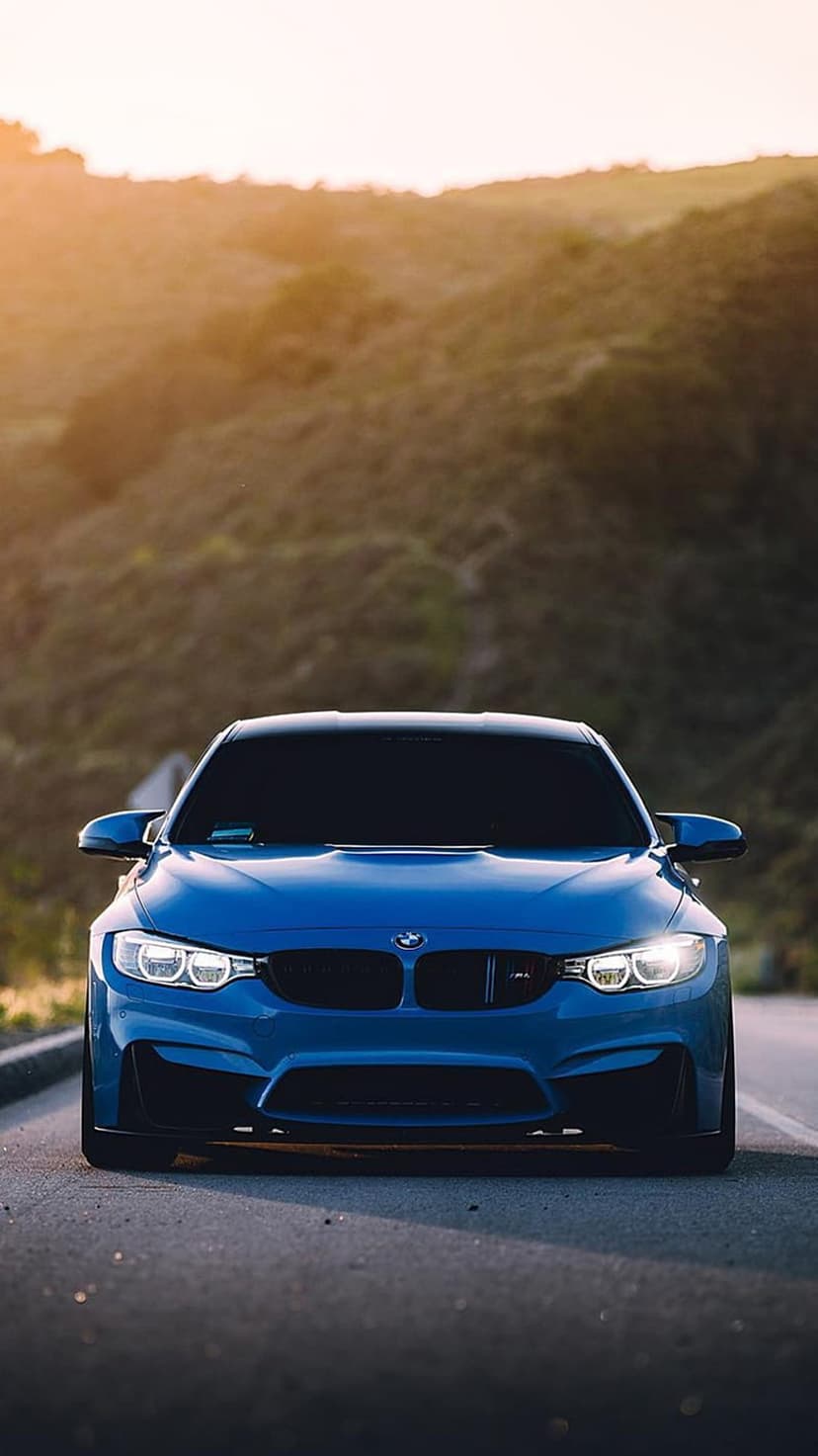 Blue BMW M4 sports car parked on a mountain road at sunset with glowing headlights and rolling hills behind it.