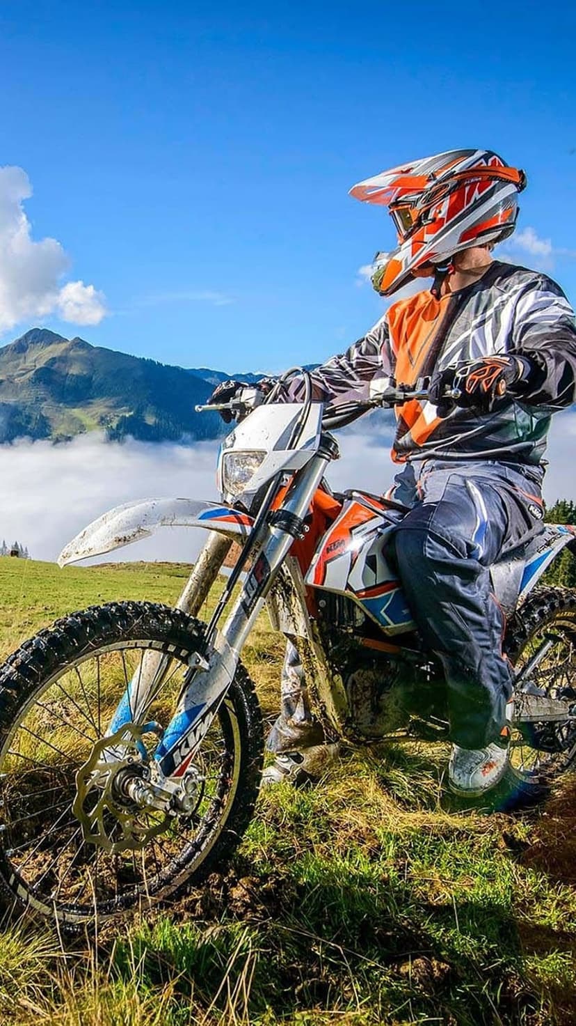 Motocross rider in full gear sits on a dirt bike atop a rocky mountain peak against a clear blue sky.