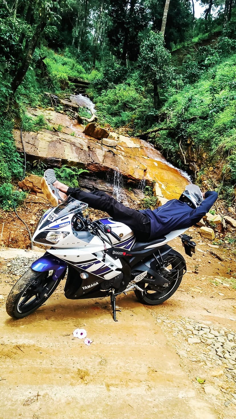 Biker in blue gear rests on a white motorcycle parked by a rocky woodland waterfall amidst dense green ferns.