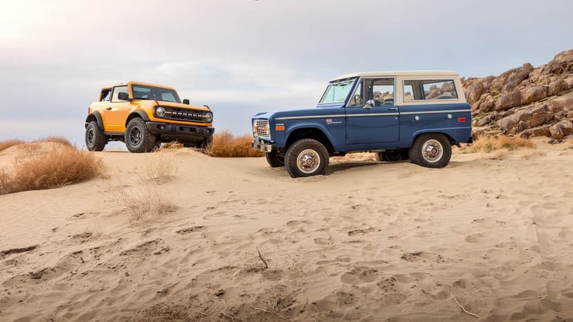 Modern yellow and classic blue Ford Broncos parked on rippled sand dunes under a soft overcast sky.