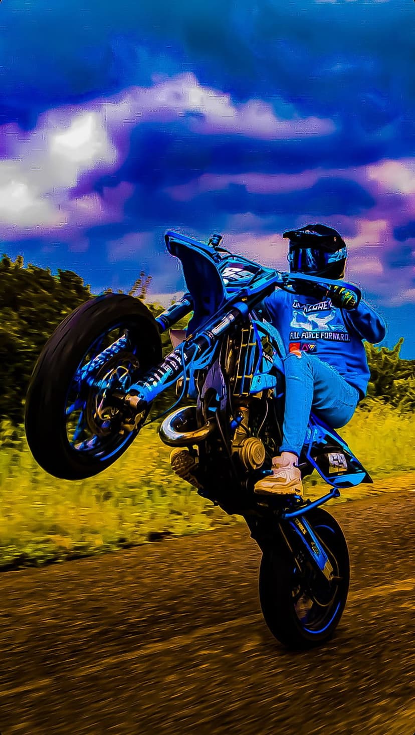 Motorcyclist in blue gear performs a wheelie on a dirt trail against dark purple and blue storm clouds.