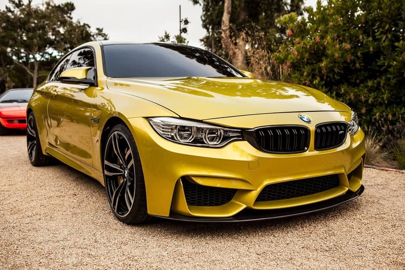 Austin Yellow BMW M4 parked on a gravel driveway surrounded by green foliage and a red car in the distance.