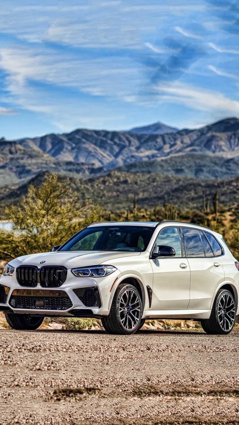 White BMW X5 M luxury SUV parked on a desert dirt road with rugged mountains under a blue sky background.