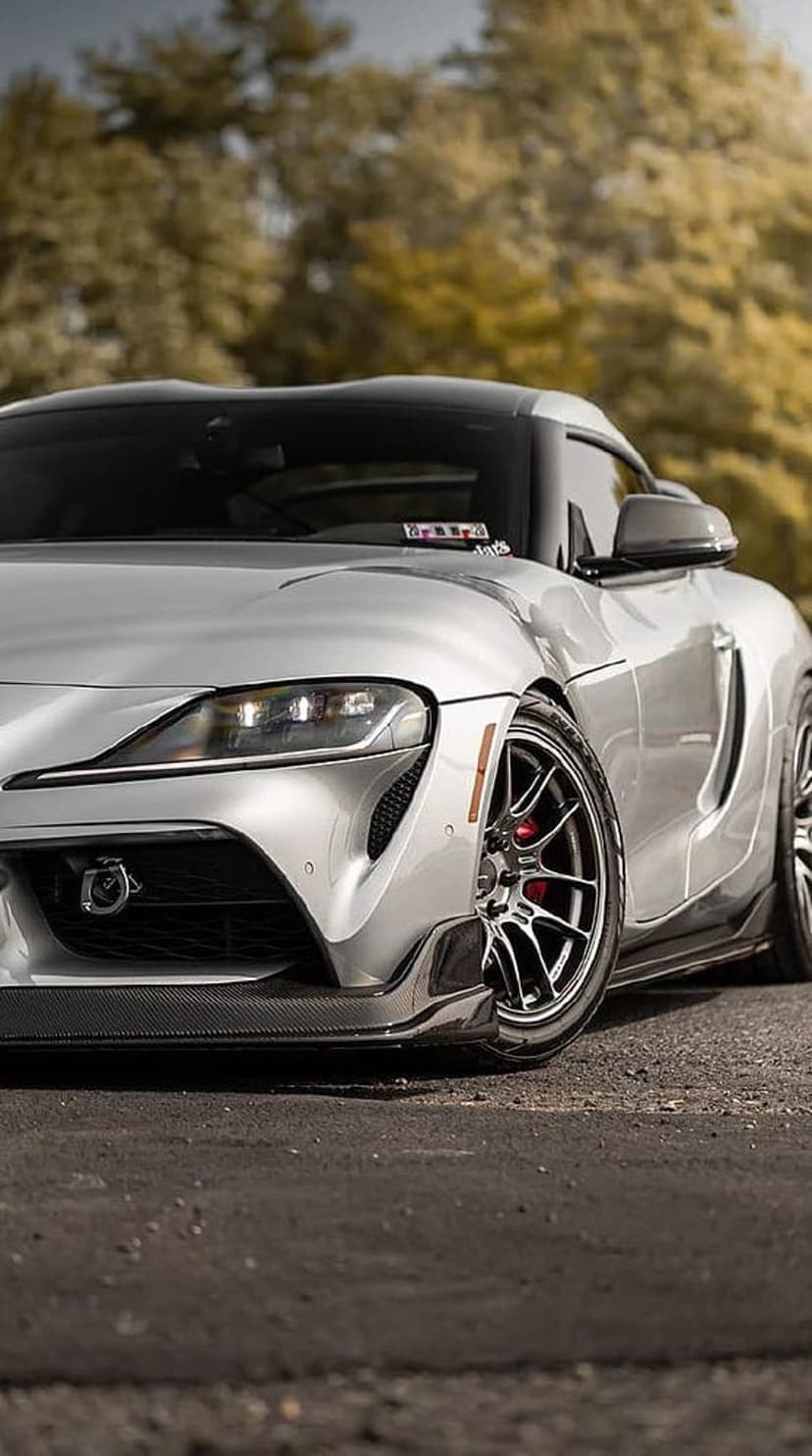 Silver sports car with red brake calipers and custom wheels reflects autumn trees in its glossy metallic paint.