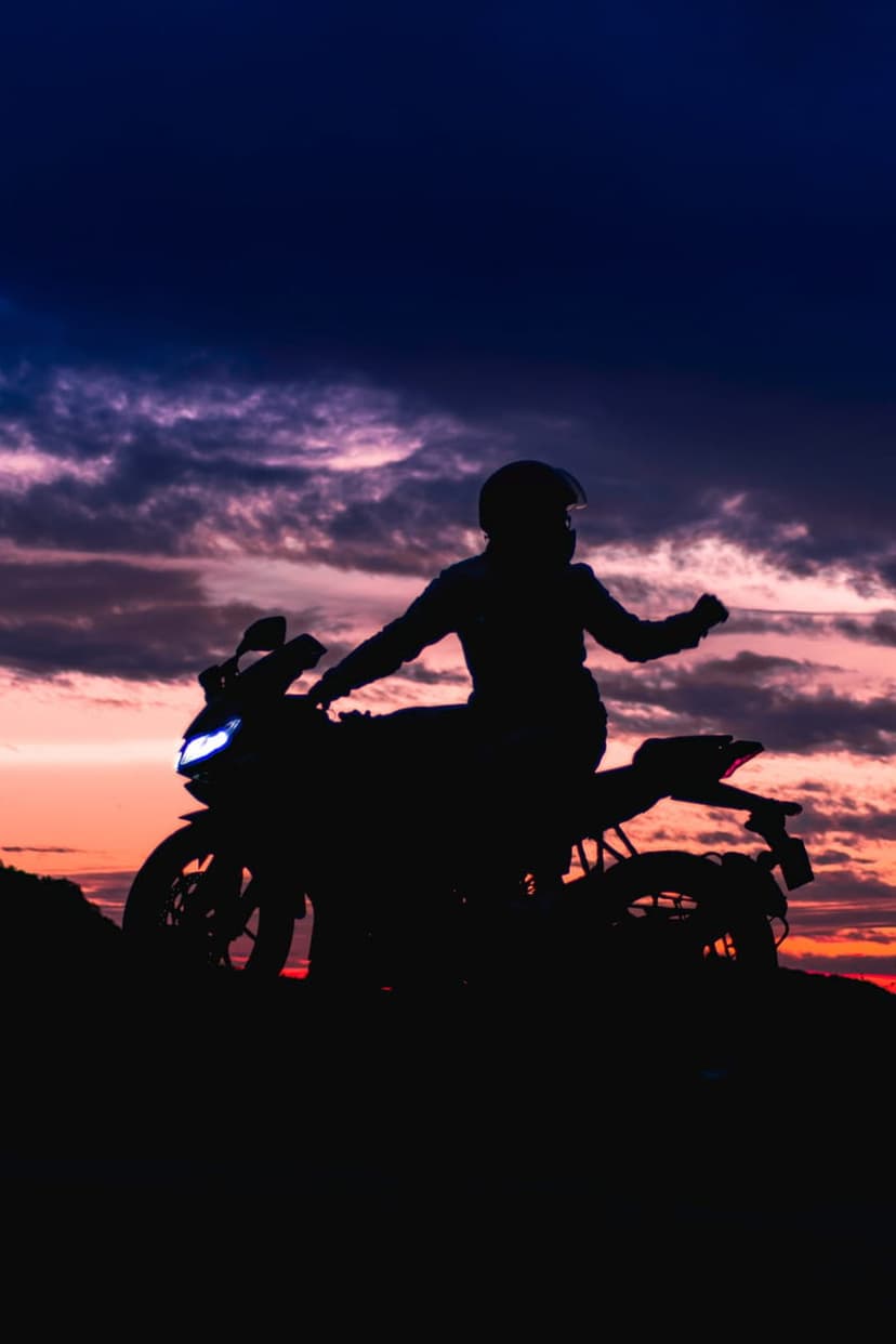 Biker silhouette stands with a motorcycle against a purple and blue sunset sky with glowing headlight beams.