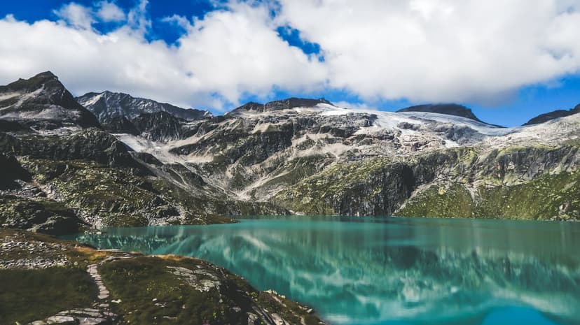 Turquoise alpine lake reflecting snow-capped mountain peaks under a blue sky with soft white cumulus clouds.