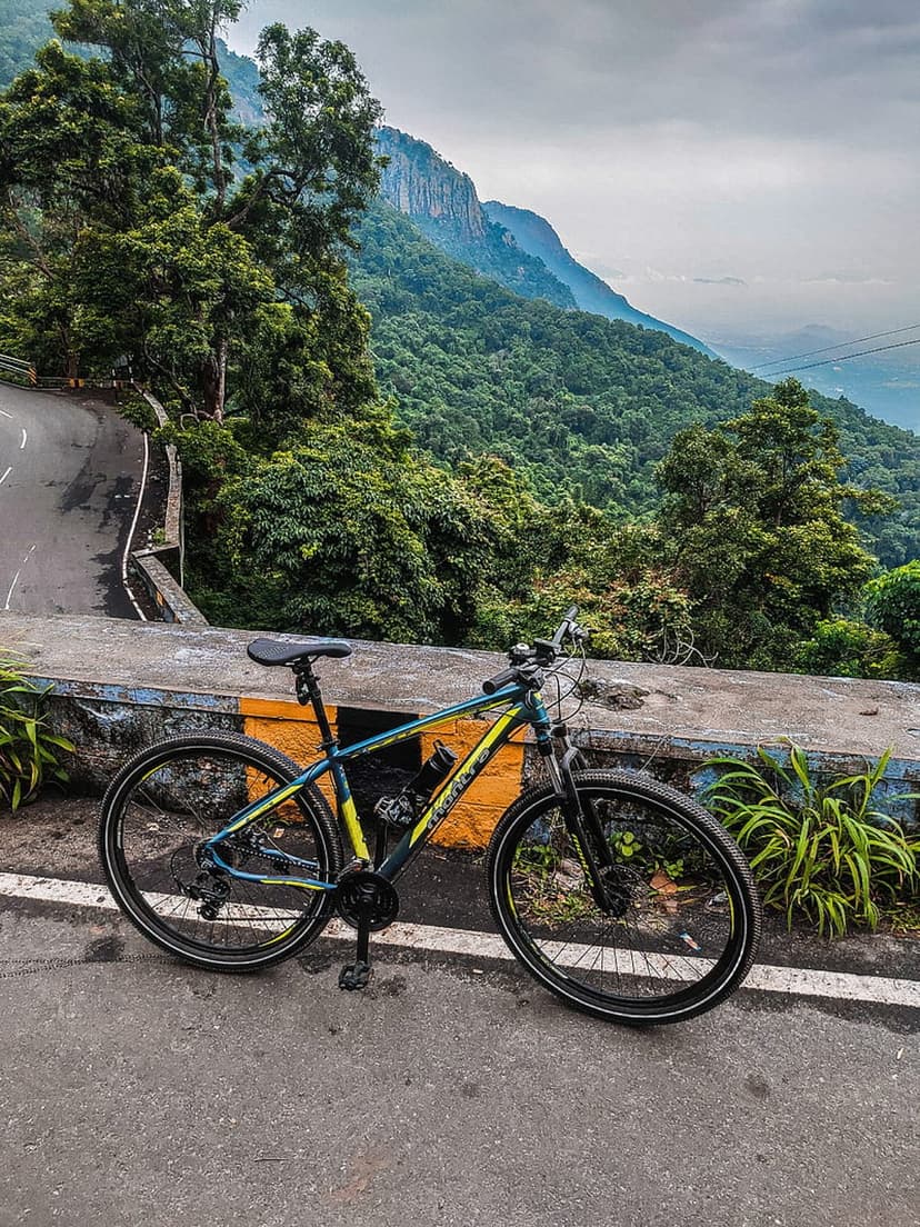Mountain bike rests by a winding road overlooking a lush forest valley and hazy peaks under a cloudy sky.