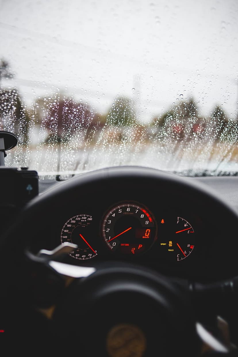 Car dashboard with glowing red gauges viewed through a windshield covered in rain droplets and gray mist.