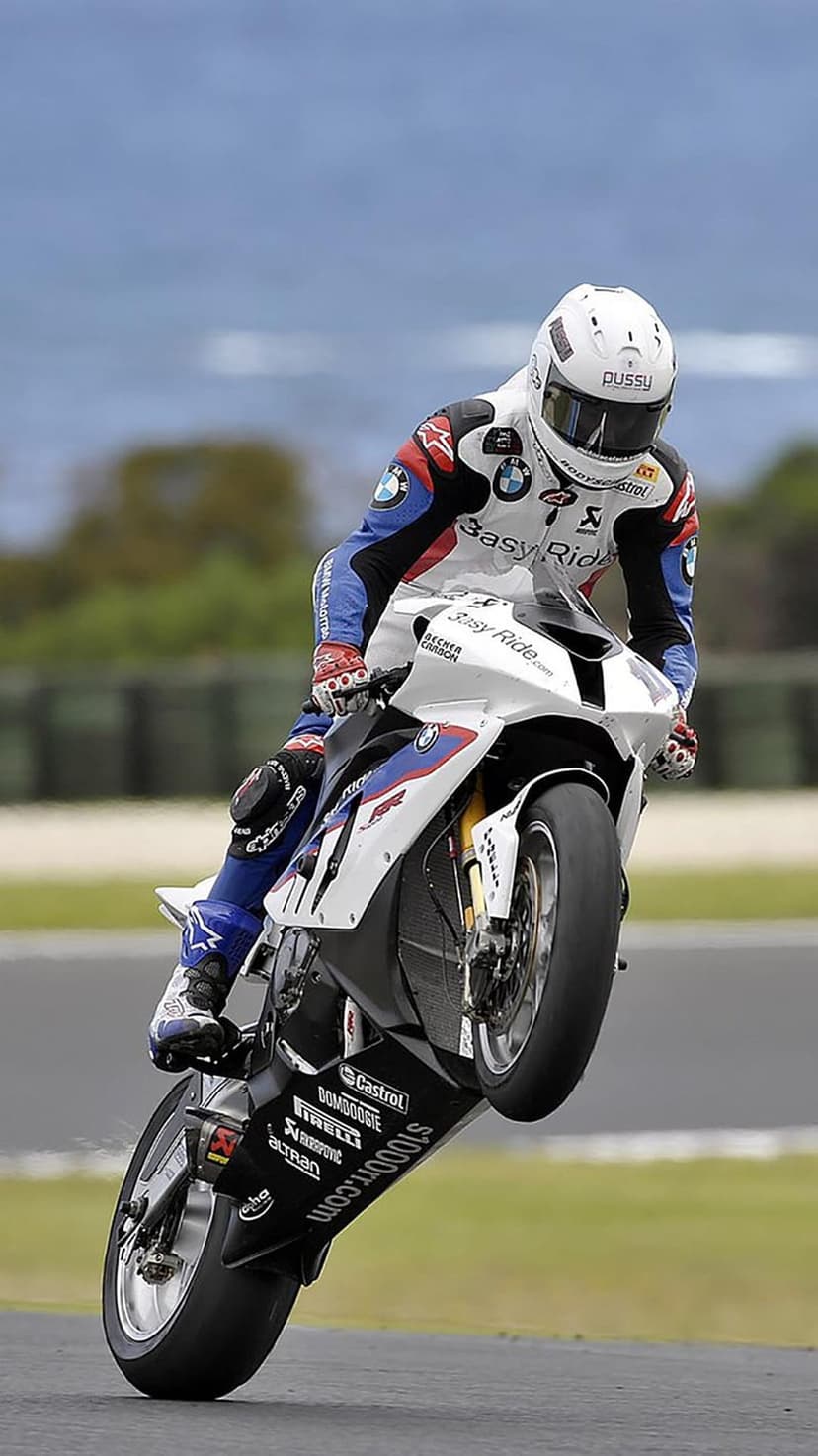 Motorcycle racer performing a wheelie on a white BMW S1000RR superbike during a high-speed track race event.