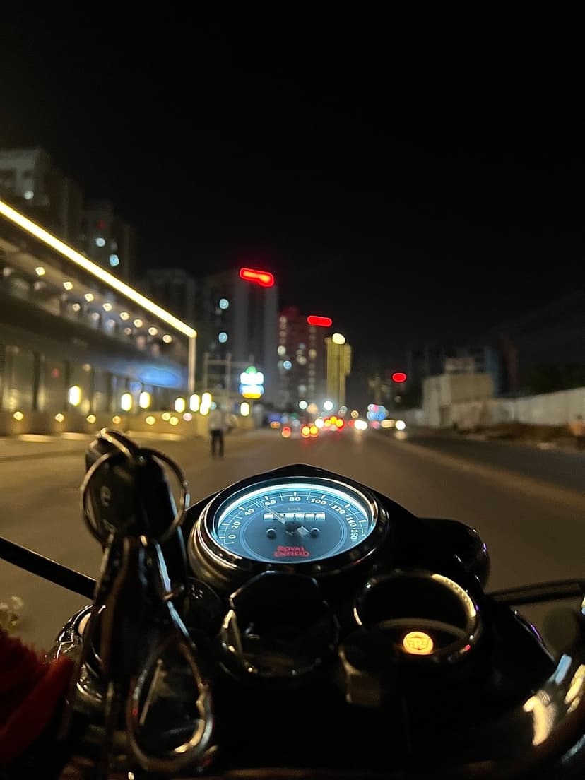Royal Enfield motorcycle speedometer with glowing orange needles against a blurred nighttime city street backdrop.