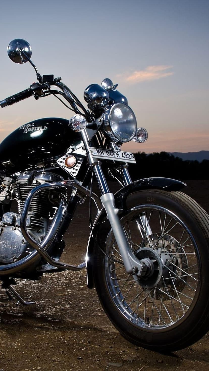 Royal Enfield motorcycle with chrome accents parked on dusty ground during a warm orange and purple sunset.