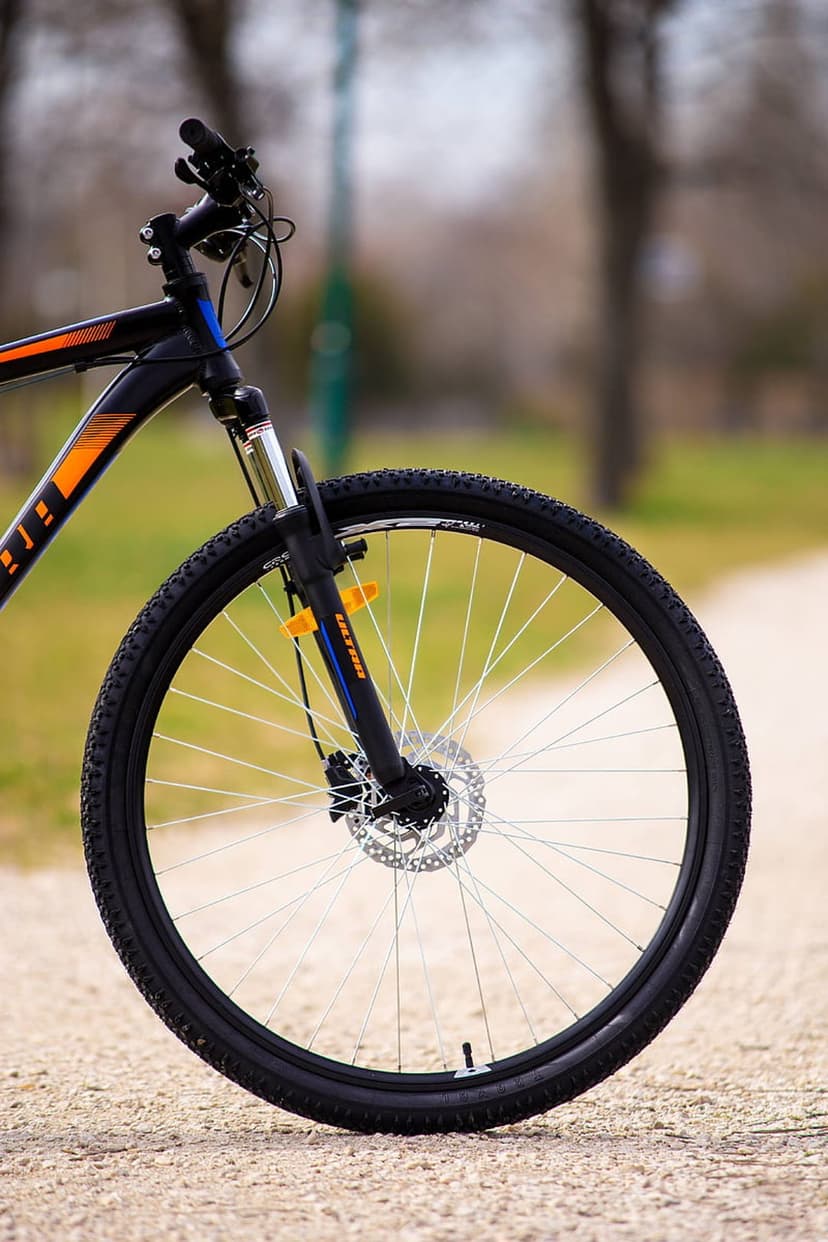 Mountain bike front wheel with an orange suspension fork and disc brake against a blurred forest path background.