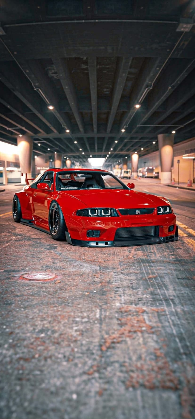 Red Nissan Skyline R32 GT-R with widebody kit parked on a concrete road under an industrial overpass at night.