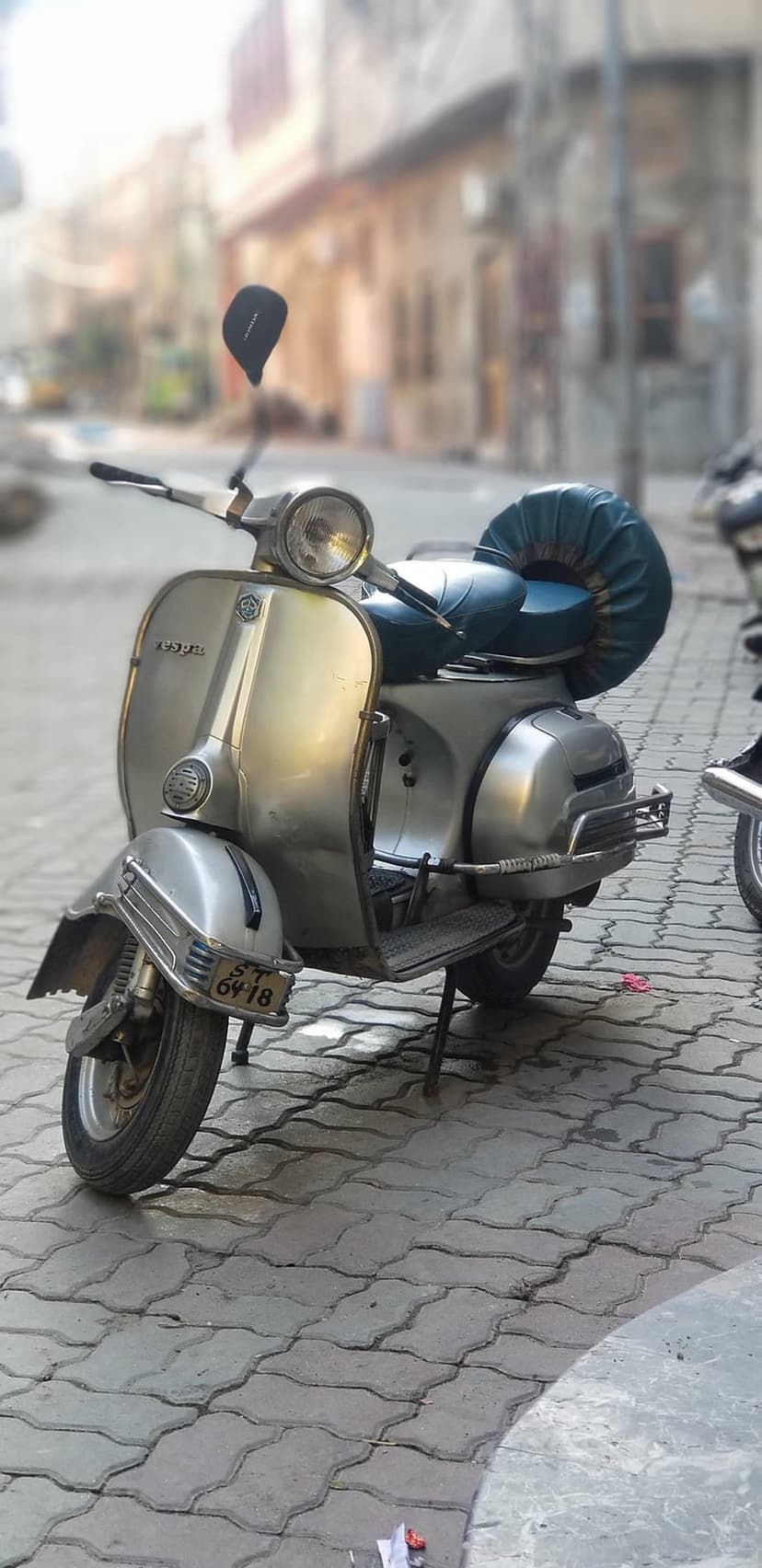 Vintage silver Vespa scooter with a teal seat parked on a textured gray cobblestone street under soft light.